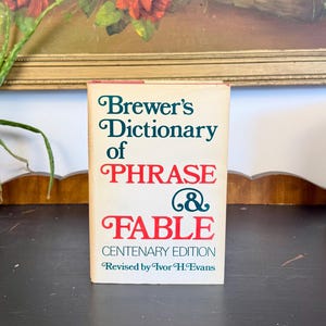 May include: A vintage copy of "Brewer's Dictionary of Phrase & Fable" with a cream-colored cover and text in green and red. The book is a centenary edition, revised by Ivor H. Evans. The book is on a black surface.