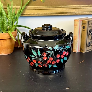 May include: A black ceramic jar with a lid, featuring a cherry design with red cherries and green leaves. The jar has two handles and sits on a dark surface. A painting of red roses is in the background.