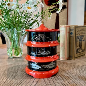 May include: A tiered, cylindrical container shaped like a pagoda, featuring a glossy red finish on the rims and a black body decorated with intricate gold-toned landscape illustrations of buildings, trees, and mountains, topped with a small finial.