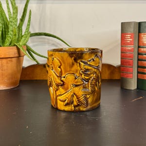 May include: A cylindrical, glazed ceramic vase in shades of brown and yellow, featuring a relief design of bees and foliage. The vase is displayed on a dark wooden surface, with a potted plant and books in the background.