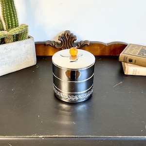 May include: A silver-colored, tiered container with a decorative leaf pattern around the base and a small, orange-colored knob on the lid. The container sits on a black surface, with a cactus in a concrete planter and books in the background.