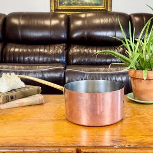 May include: A round copper saucepan with a long, straight brass handle and a silver-toned interior. The exterior of the pan features a smooth, polished copper finish, while the handle is securely attached to the side of the vessel.