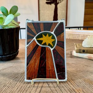 May include: A rectangular stained glass panel featuring a central yellow star set against a dark green circular background. The design includes radiating orange and dark red sunburst rays with intricate black patterns, all encased in a silver metal frame.