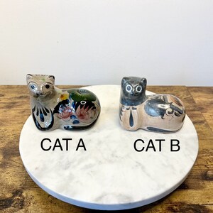May include: Two ceramic cat figurines labeled CAT A and CAT B. CAT A features a light base with green, blue, and pink floral patterns. CAT B has a tan base with black and orange painted details. Both figures are in a resting, seated position.