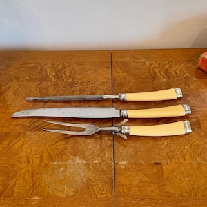 May include: A vintage carving set with three pieces: a knife sharpener, a carving knife, and a carving fork. The handles are a light cream color with silver-toned accents. The set is displayed on a wooden surface.