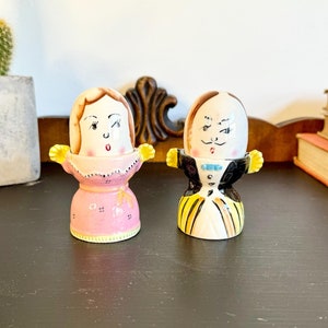 May include: A pair of vintage ceramic egg cups, one depicting a woman in a pink dress and the other a man in a tuxedo. The egg cups have hand-painted details and are displayed on a dark surface.
