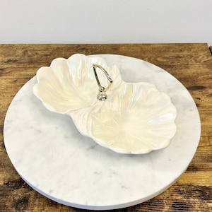 May include: A two-sectioned, iridescent white serving dish with a silver handle, resting on a round, white marble tray. The dish has a scalloped edge, and the tray sits on a wooden surface.