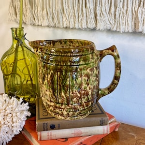 May include: A vintage ceramic pitcher with a handle, featuring a brown and green pattern, sits atop a stack of old books. A green glass bottle with a plant and a piece of white coral are also on the table.