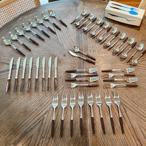 May include: A large stainless steel flatware set featuring dark brown handles, including various spoons, forks, and knives arranged on a wooden table. A white rectangular box with blue graphic illustrations of the utensils is included in the set.