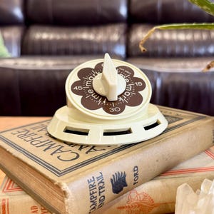 May include: A cream-colored kitchen timer with a brown floral-shaped dial featuring the text "time mark" and numbers 15, 30, and 45. The timer has a central knob and sits atop two stacked vintage books titled "Campfire and Trail" by Ford.