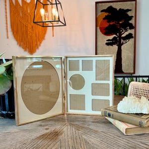 May include: A hinged double photo frame with a metallic gold finish. The left side features a large oval mat opening, while the right side contains a multi-opening mat with one circular and five rectangular cutouts for displaying multiple photographs.
