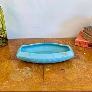 May include: A light blue ceramic dish with a textured surface. The dish is oval-shaped and sits on a wooden table. A green glass bottle and books are in the background.