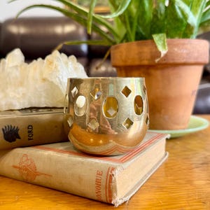 May include: A gold-toned metal votive candle holder featuring a repeating pattern of circular and diamond-shaped cutouts. The cylindrical object has a smooth, reflective surface and sits atop a stack of two vintage books with visible text.