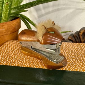 May include: A wooden stapler with a lion head design, featuring a brown body and a faux fur mane. The stapler rests on a patterned surface atop a stack of books. An aloe vera plant in a terracotta pot is in the background.