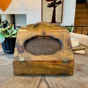 May include: A wooden bread box with a medium brown finish, featuring a hinged front door with a central cutout window. The door is secured by a small round knob and two brass-colored metal hinges at the base, showcasing a natural wood grain.