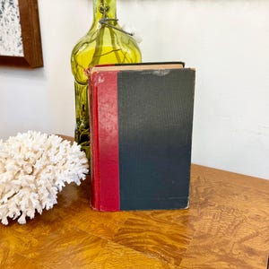 May include: A still life arrangement featuring a vintage book with a red spine and black cover, a green glass bottle, and a white coral piece. The items are arranged on a wooden surface, creating a rustic aesthetic.
