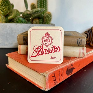 May include: A vintage Stroh's Beer coaster with a red and white logo featuring a lion and crown. The coaster rests on a stack of old books with orange covers, next to a potted cactus.
