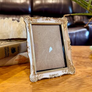 May include: A rectangular picture frame with an off-white finish and ornate gold-toned metallic detailing along the edges. The frame features intricate scrollwork patterns at the corners and sides, surrounding a plain, neutral-colored insert.