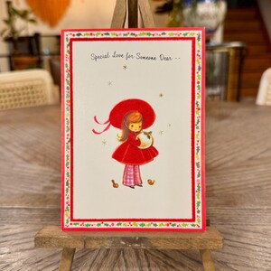 May include: A vintage greeting card with a red-framed border and the text "Special Love for Someone Dear --". The card features an illustration of a girl in a red dress and hat, holding a basket. The card is displayed on a small wooden easel.