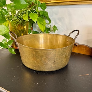 May include: A vintage brass bowl with two small handles. The bowl has a wide, shallow design and a slightly tarnished finish, suggesting age. The bowl is sitting on a black surface, with a green plant in the background.