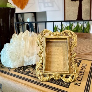 May include: A small, rectangular, gold-colored metal picture frame featuring an ornate, scrolling leaf-like border and a beaded inner edge. The frame is empty, revealing a plain, light brown backing material within the central display area.