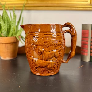May include: A brown ceramic pitcher with a handle. The pitcher features a relief design of dogs in a field. The pitcher is sitting on a dark surface, with a plant and framed artwork in the background.