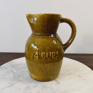 May include: A small, brown ceramic pitcher with a handle, labeled "4 CUPS" in white lettering. The pitcher has a speckled texture and a glossy finish. It is sitting on a white marble surface.
