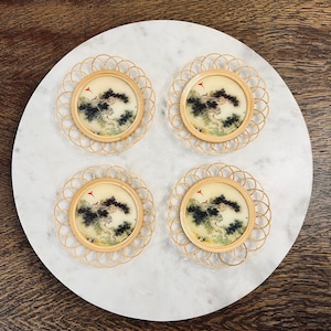 May include: Four round coasters with a bamboo woven border. Each coaster features a circular image of a tree and bird scene. The coasters are arranged on a white marble surface, set against a dark wood background.