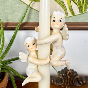 May include: A cream-colored candle with two ceramic cherub figurines. The cherubs have white wings and are hugging the candle. The background includes a green and brown embroidered design and a potted plant.