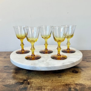 May include: Set of five amber-colored wine glasses. Each glass features a clear bowl, a stem with a decorative design, and a round base. The glasses are arranged on a white marble serving tray, showcasing their vintage aesthetic.
