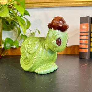 May include: A ceramic turtle figurine in shades of green with a brown hat. The turtle has a speckled texture and decorative swirls on its shell. The figurine is a decorative item.