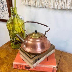 May include: A copper tea kettle with a brass lid and handle sits atop two vintage books with orange covers. A green glass bottle with a white stopper is in the background. The kettle has a dark brown handle.