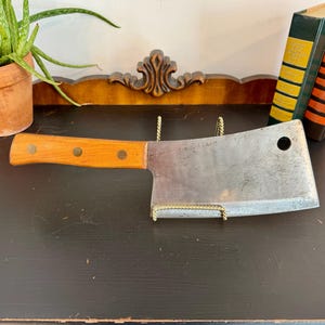 May include: A vintage cleaver with a wooden handle and a large, rectangular steel blade. The handle is light brown with three rivets. The cleaver is displayed on a black surface with a decorative wooden accent and two books.