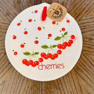 May include: Round white pot holder with a red loop and cherry design. The pot holder features red cherries with green leaves and the word "cherries" in red. A brown tag is attached.