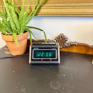 May include: A black Bulova digital clock displaying 12:06 PM with a green LED display. The clock has a silver-colored trim and sits on a dark surface. A potted aloe vera plant and a painting with red flowers are in the background.