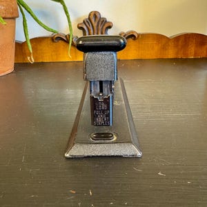 May include: A vintage-style black and dark gray metal stapler with a smooth top handle. The front vertical plate features white text that reads: TO LOAD PULL UP AND INSERT STAPLES. The base is wide and rectangular with a textured finish and a small black button.