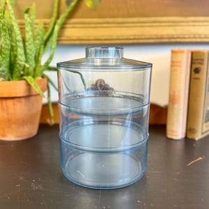 May include: A clear, three-tiered serving dish with a light blue tint. The dish has a lid and three circular trays. The container is made of a transparent material, allowing visibility of the contents. The dish is suitable for serving snacks or desserts.