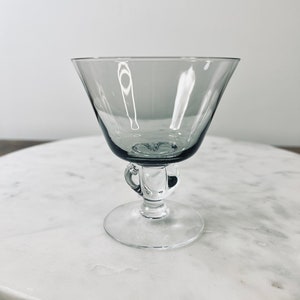 May include: A gray-tinted glass cocktail coupe with a flared bowl, a small handle, and a round base. The stem features a decorative bulbous design. The glass is transparent, allowing light to pass through. The coupe is designed for serving cocktails.