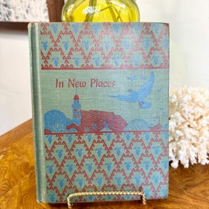 May include: A hardcover book titled In New Places with a light green cover featuring a red and blue geometric pattern, a red building illustration, and a blue bird in flight. The book is displayed upright on a gold-toned metal stand.