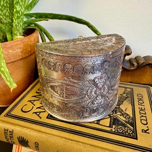 May include: A silver-colored, antique, half-moon-shaped box with intricate embossed floral and architectural designs. The box is resting on a vintage book with the title "Campfire and Trail" and is next to a potted aloe vera plant.