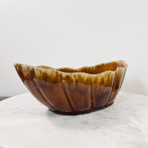 May include: A brown ceramic bowl with a scalloped rim. The bowl has a glossy finish and a dark brown color that fades to a lighter brown at the rim. The bowl is sitting on a white marble surface.