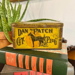 May include: A vintage rectangular tin with the words "DAN PATCH CUT PLUG" in black and gold lettering. The tin features an illustration of a horse-drawn carriage. The tin is resting on top of a stack of old books.