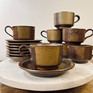 May include: A set of vintage ceramic cups and saucers. The cups and saucers are brown with a textured, geometric design. The cups have a rounded shape with a handle. The saucers are round and flat. The set is displayed on a white marble surface.