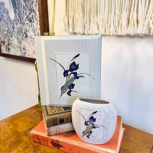 May include: A white ceramic vase and a matching square wall art piece, both featuring a blue iris floral design. The vase is oval-shaped and sits atop a stack of vintage books. The wall art has a striped background.