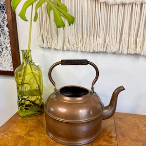 May include: A vintage copper kettle with a dark wooden handle and spout. The kettle is round with a slightly flattened base. A green glass bottle with a plant is to the left of the kettle. A macrame wall hanging is in the background.