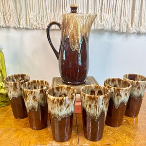 May include: A ceramic beverage set consisting of a tall pitcher with a lid and six matching tumblers. Each piece features a glossy dark brown base glaze with a cream-colored drip pattern cascading from the rim down the sides of the vessels.