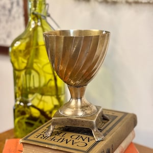 May include: A vintage brass chalice with a spiral design, resting on a stack of old books. The chalice has a square base with decorative feet. A green glass bottle is in the background.