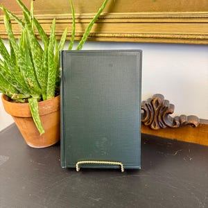 May include: A dark green hardcover book with a simple embossed border design on the front cover. The book is displayed upright on a small, gold-toned metal wire stand, showcasing its solid-colored binding and clean, minimalist front cover.