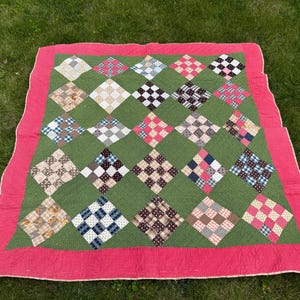 May include: A handmade quilt with a green background and a red border. The quilt features a diamond pattern with various fabric designs in shades of brown, blue, pink, and white. The quilt is displayed on a bed of green grass.