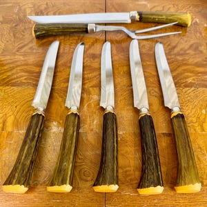 May include: A set of five steak knives and one carving fork with silver-toned metal blades and tines, featuring textured, dark brown, antler-style handles with yellow-toned end caps, arranged on a polished wooden surface with a visible grain pattern.